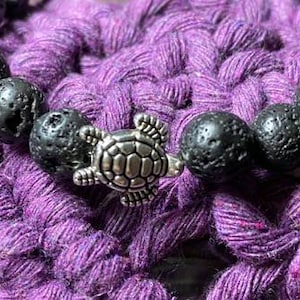 May include: A black lava rock bracelet with a silver turtle charm. The bracelet is on a purple crocheted surface.