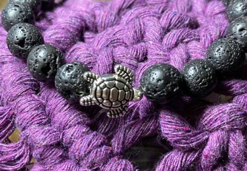 May include: A black lava rock bracelet with a silver turtle charm. The bracelet is on a purple crocheted surface.