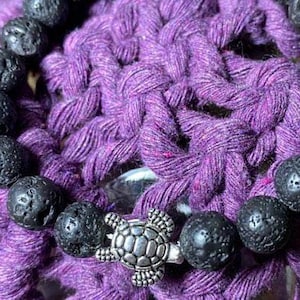 May include: A black lava rock bracelet with a silver turtle charm. The bracelet is on a purple crocheted surface.
