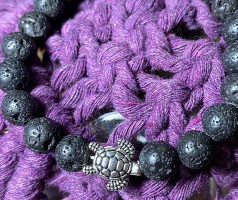 May include: A black lava rock bracelet with a silver turtle charm. The bracelet is on a purple crocheted surface.