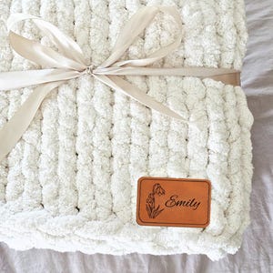 May include: A chunky knit cream-colored throw blanket made of thick, soft yarn. The blanket is folded and tied with a smooth, light beige satin ribbon bow. A rectangular tan leather patch is attached to the corner, featuring a floral icon and the name Emily.
