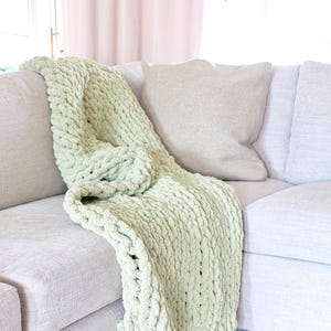 Puede incluir: Una manta de punto grueso verde claro sobre un sof&aacute; gris claro. La manta tiene un tejido grueso y texturizado y est&aacute; doblada sobre el brazo del sof&aacute;. Una almohada beige descansa contra el respaldo del sof&aacute;.