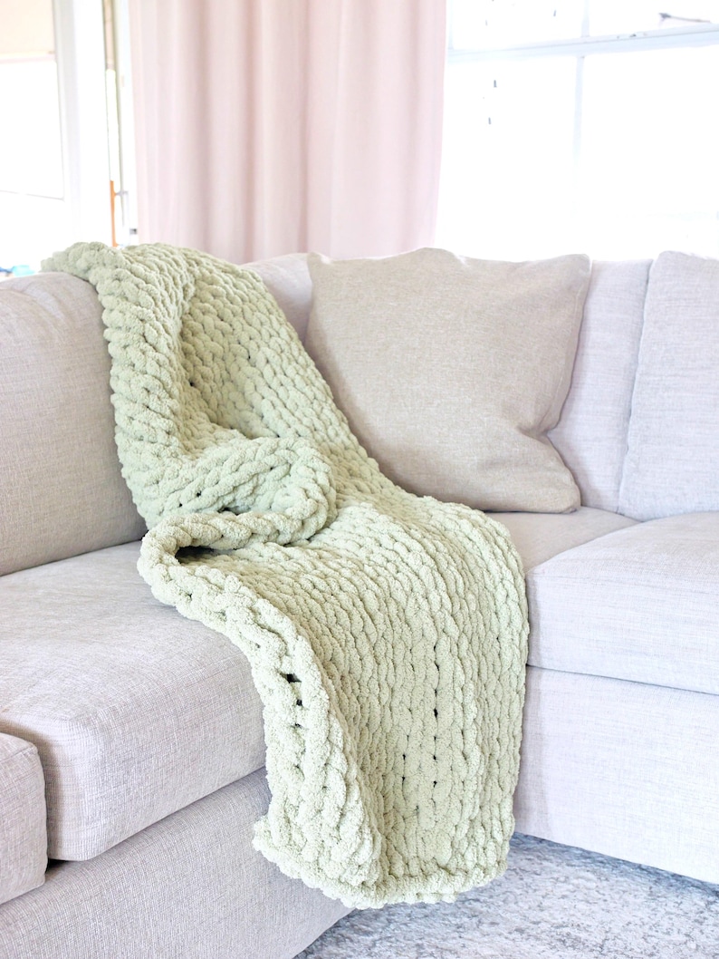 Puede incluir: Una manta de punto grueso verde claro sobre un sof&aacute; gris claro. La manta tiene un tejido grueso y texturizado y est&aacute; doblada sobre el brazo del sof&aacute;. Una almohada beige descansa contra el respaldo del sof&aacute;.