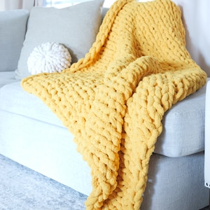 Puede incluir: Una manta de punto grueso de color amarillo mostaza sobre un sof&aacute; gris. La gruesa textura de la manta es visible, con un coj&iacute;n a juego y un coj&iacute;n decorativo blanco. La escena est&aacute; ambientada en un sal&oacute;n bien iluminado.