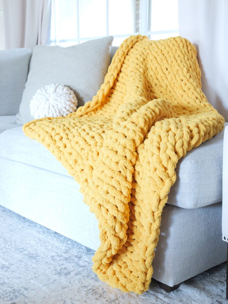 Puede incluir: Una manta de punto grueso de color amarillo mostaza sobre un sof&aacute; gris. La gruesa textura de la manta es visible, con un coj&iacute;n a juego y un coj&iacute;n decorativo blanco. La escena est&aacute; ambientada en un sal&oacute;n bien iluminado.