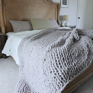 Puede incluir: Una manta de punto grueso gris claro sobre una cama. La cama tiene un cabecero de madera y est&aacute; hecha con s&aacute;banas blancas y almohadas grises. La textura de la manta es visible, lo que sugiere una sensaci&oacute;n suave y acogedora. La habitaci&oacute;n tiene una paleta de colores neutros.