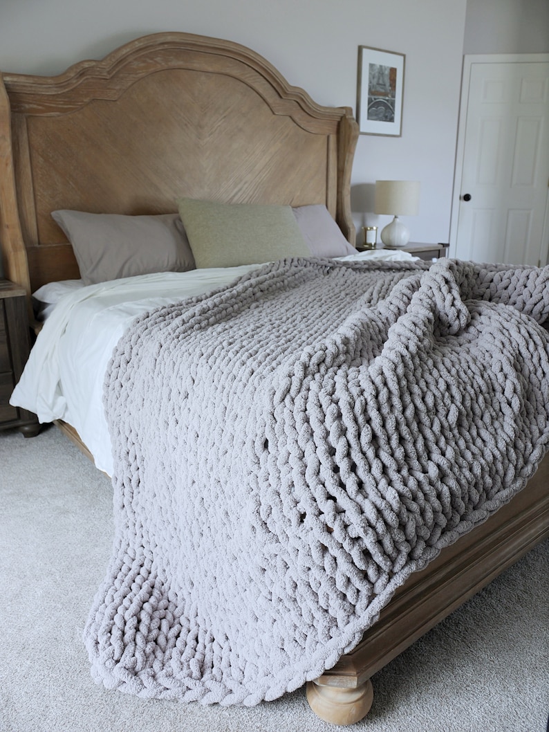 Puede incluir: Una manta de punto grueso gris claro sobre una cama. La cama tiene un cabecero de madera y est&aacute; hecha con s&aacute;banas blancas y almohadas grises. La textura de la manta es visible, lo que sugiere una sensaci&oacute;n suave y acogedora. La habitaci&oacute;n tiene una paleta de colores neutros.