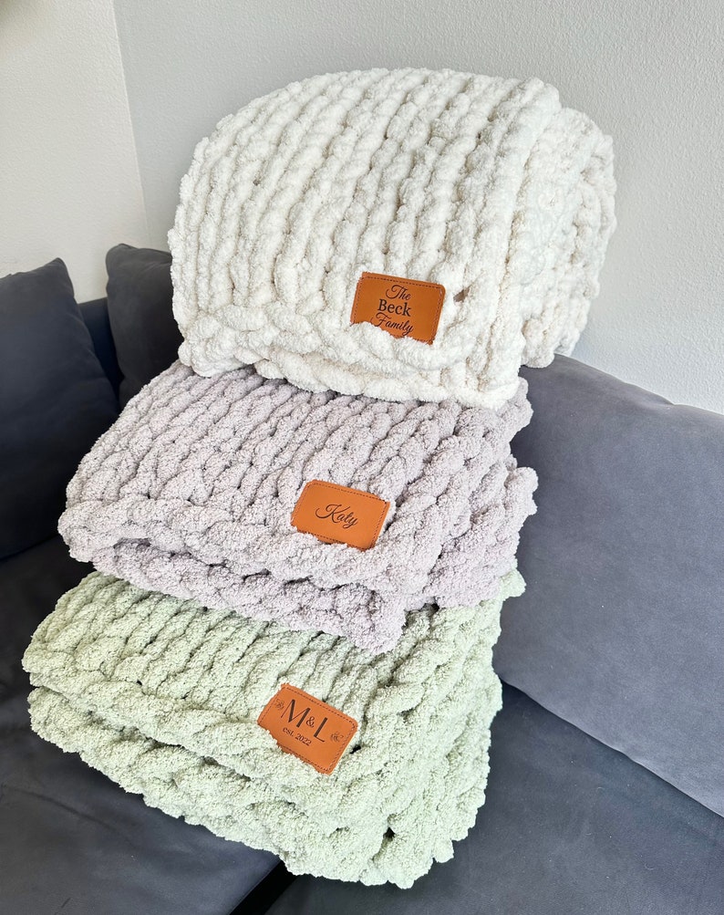 Puede incluir: Tres mantas de punto grueso en crema, lavanda y verde salvia apiladas. Cada manta tiene un parche de cuero marr&oacute;n con texto personalizado: "The Beck Family", "Koty" y "M&L Est. 2022".