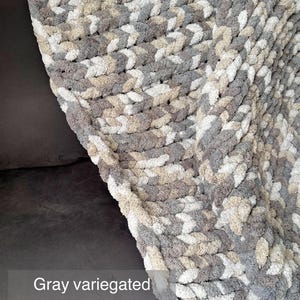 Puede incluir: Una manta de punto grueso gris jaspeado. La manta est&aacute; hecha de hilo grueso y suave en tonos de gris, beige y blanco. El texto "Gray variegated" es visible en la imagen. La manta est&aacute; sobre una superficie gris oscuro.