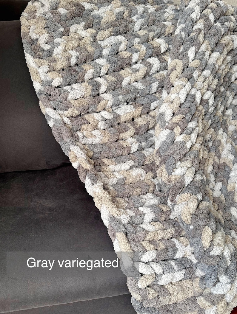Puede incluir: Una manta de punto grueso gris jaspeado. La manta est&aacute; hecha de hilo grueso y suave en tonos de gris, beige y blanco. El texto "Gray variegated" es visible en la imagen. La manta est&aacute; sobre una superficie gris oscuro.