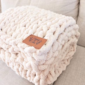 May include: A chunky knit blanket in a soft cream color with a thick, braided texture. A rectangular brown leather patch is stitched onto the front, featuring the text Isabella & John with a small botanical illustration of leaves on each side.
