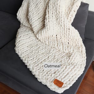 Puede incluir: Una manta de punto grueso de color crema sobre un sof&aacute; gris oscuro. La manta tiene un aspecto grueso y texturizado con la palabra "Oatmeal" impresa. Una peque&ntilde;a etiqueta de cuero marr&oacute;n est&aacute; adherida a la manta.