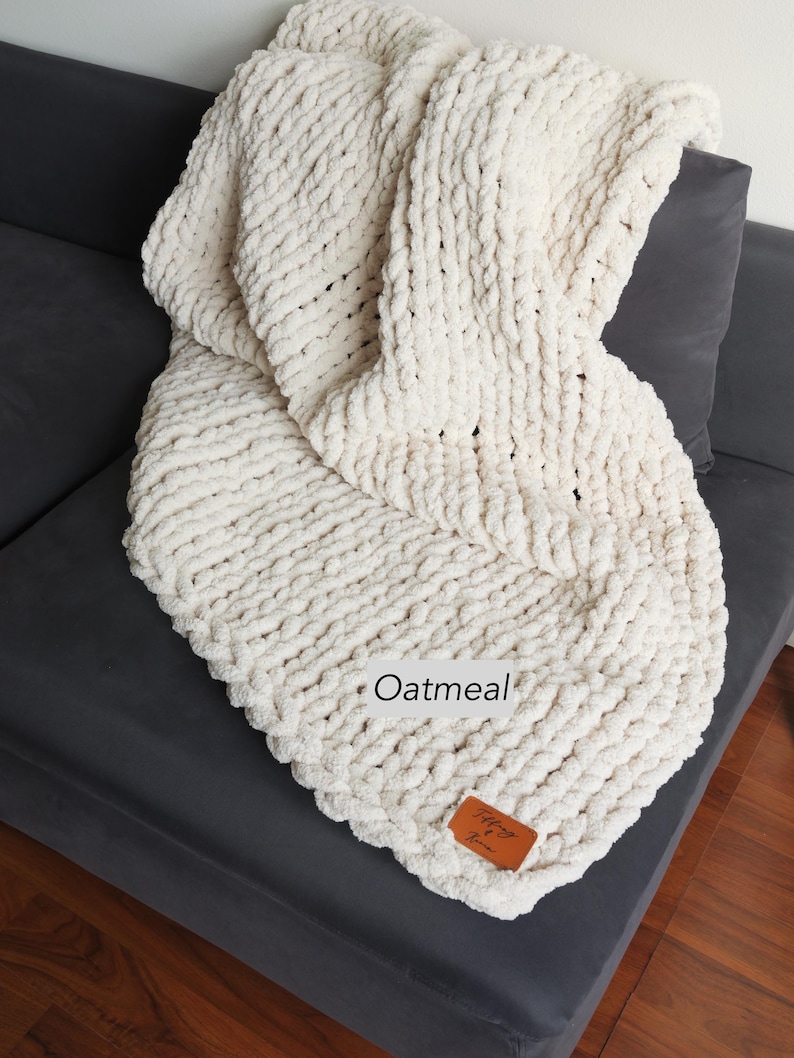 Puede incluir: Una manta de punto grueso de color crema sobre un sof&aacute; gris oscuro. La manta tiene un aspecto grueso y texturizado con la palabra "Oatmeal" impresa. Una peque&ntilde;a etiqueta de cuero marr&oacute;n est&aacute; adherida a la manta.