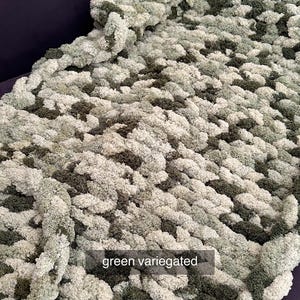 Puede incluir: Una manta de punto grueso en tonos verdes, blancos y verde oscuro. La manta tiene un aspecto texturizado y afelpado. El texto "green variegated" se muestra en la manta.