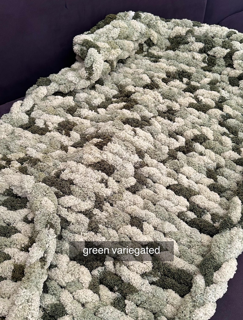 Puede incluir: Una manta de punto grueso en tonos verdes, blancos y verde oscuro. La manta tiene un aspecto texturizado y afelpado. El texto "green variegated" se muestra en la manta.