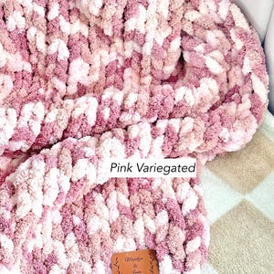 Puede incluir: Una manta de punto grueso en un esquema de color rosa variado. La manta presenta una mezcla de tonos rosas claros y oscuros, creando una apariencia texturizada. Una peque&ntilde;a etiqueta de cuero marr&oacute;n con el texto "Maritza & Joan" est&aacute; adjunta.