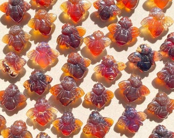 May include: A collection of amber bee charms in various shades of orange, brown, and red. The charms are carved in a detailed bee shape with wings and legs.