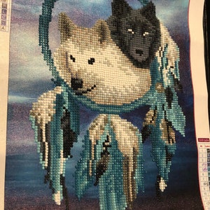 May include: A diamond painting kit depicting two wolves inside a dreamcatcher. The dreamcatcher is blue and white with feathers. The wolves are white and black. The background is a dark blue with stars. The kit includes a drill table with numbers and symbols.