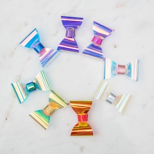 May include: Assortment of iridescent bow hair clips in various colors, including blue, purple, green, gold, and red. The bows are arranged in a circle on a white marble surface.