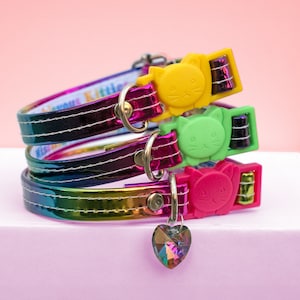 May include: Three cat collars stacked on top of each other. The collars are iridescent with a rainbow sheen. Each collar has a different colored buckle: yellow, green, and pink. A heart-shaped charm hangs from the bottom collar.