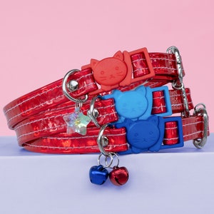 May include: Three red cat collars with a silver buckle and a cat-shaped charm. The collars are stacked on top of each other. The top collar has a red cat charm, the middle collar has a blue cat charm, and the bottom collar has a blue cat charm. The collars are decorated with a silver star charm and two silver bells.