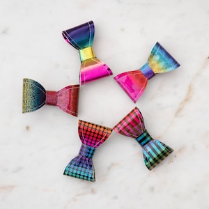 May include: Five colorful bow-shaped hair clips with different patterns. The bows are made of iridescent material and have a variety of colors, including pink, blue, green, and purple.