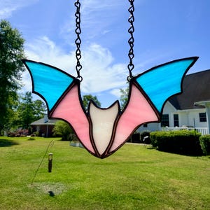 May include: A stained glass bat with pink, white, and blue wings hangs from a black chain. The bat is suspended against a backdrop of a bright blue sky and green grass.