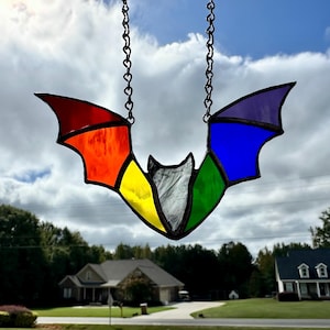 Peut inclure: Un vitrail en forme de chauve-souris, suspendu par une chaîne noire, avec des ailes aux couleurs de l'arc-en-ciel. Les ailes de la chauve-souris sont segmentées en rouge, orange, jaune, vert, bleu et violet. Le fond montre un ciel nuageux et des maisons.
