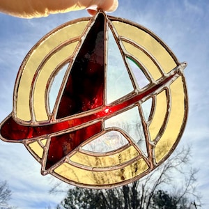 Puede incluir: Un emblema de vidriera con un anillo exterior amarillo, una franja roja y un centro rojo oscuro y transparente. El emblema se sostiene contra un cielo azul con ramas de árboles visibles en el fondo.