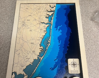 May include: A wooden map of Barnegat Bay, Long Beach Island, New Jersey. The map is layered with shades of blue to represent the ocean depths. The map includes a compass and coordinates for the location. The text "Barnegat Bay" and "LBI - New Jersey" is engraved on the map.