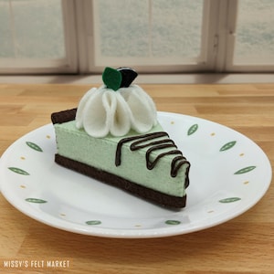 May include: A felt mint chocolate chip pie slice with a chocolate drizzle on a white plate with a green leaf pattern.