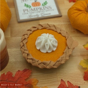 May include: A felt pumpkin pie with a brown crust and a white whipped cream topping. The pie is on a wooden surface with fall leaves.