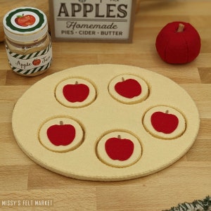 May include: A felt craft featuring a light beige circular base with five red apple cutouts. A jar of apple jam and a felt apple are also visible. The sign in the background reads "APPLES Homemade PIES CIDER BUTTER".