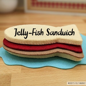 May include: A felt play food sandwich shaped like a jellyfish. The sandwich is made of three layers of felt: a light brown layer, a red layer, and a light brown layer. The top layer has the words "Jelly-Fish Sandwich" written in black.