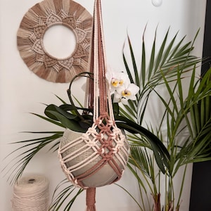 Può includere: Un portavaso in macramè beige e marrone, che tiene un vaso di ceramica grigio con una pianta verde e fiori di orchidea bianchi. Un arazzo da parete in macramè abbinato è visibile.
