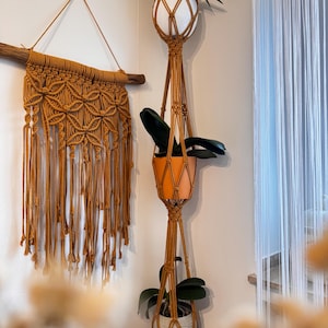 Op de afbeelding: Een macramé plantenhanger met drie potten, elk met een groene plant. De hanger is gemaakt van natuurlijke bruine koord en hangt aan het plafond. De potten zijn wit, oranje en grijs.