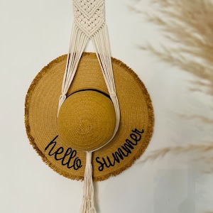 May include: A white macrame hat hanger with a wooden ring at the top. A brown straw hat with the text "hello summer" is hanging from the hanger.