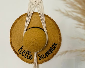 May include: A white macrame hat hanger with a wooden ring at the top. A brown straw hat with the text "hello summer" is hanging from the hanger.