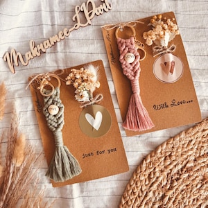 Pode incluir: Dois chaveiros de macramé, um verde sálvia e outro rosa empoeirado, cada um com uma borla e uma conta de madeira que diz "LOVE". Estão presos a um cartão de papel kraft com flores secas e um adesivo de coração. O cartão diz "just for you" e "With Love..."