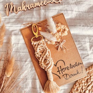 Pode incluir: Um porta-chaves de macramé bege com fecho dourado, exibido num cartão castanho com as palavras "Herzlichen Dank!" e um coração. A imagem inclui flores secas e uma placa de madeira com "Makramee L'Dart".