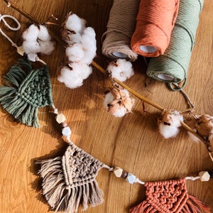 Könnte beinhalten: Handgefertigte Makramee-Wandbehänge in Grün-, Beige- und Terrakotta-Tönen mit Holzperlen. Baumwollblüten und Garnrollen in passenden Farben sind auf einer Holzoberfläche angeordnet und schaffen eine rustikale, böhmische Ästhetik.