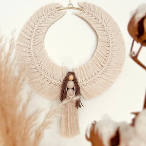 Op de afbeelding: Een beige macramé engel wandhanger met een houten poppenfiguur en een gouden hoepel. De engel heeft lang bruin haar en houdt een kleine ster vast.