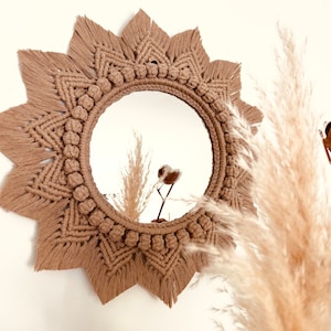 Puede incluir: Un espejo redondo de macramé color beige con diseño de sol. El marco presenta intrincados nudos y una serie de pequeños nudos decorativos alrededor del círculo interior. El espejo se muestra contra una pared blanca, con una planta borrosa en primer plano.