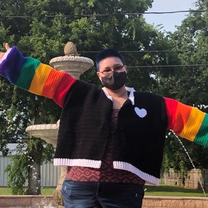 May include: A person wearing a black crochet cardigan with rainbow colored sleeves and a white heart on the front. The cardigan has a white trim around the edges. The person is wearing blue jeans and a face mask.
