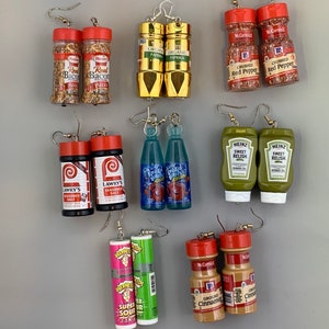 Op de afbeelding: Een verzameling novelty oorbellen gemaakt van miniatuur voedselcontainers. De oorbellen bevatten kruidenpotjes, ketchupflessen en snoepverpakkingen. De oorbellen zijn bevestigd aan zilverkleurige oorhaken.