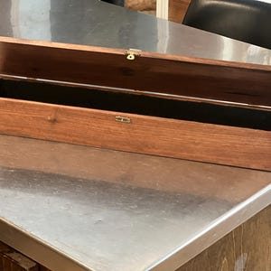 May include: A long, rectangular wooden box with a dark brown finish and visible wood grain. The box features a hinged lid that is propped open, revealing a dark interior. It is secured with a small, centered gold-toned metal latch on the front.
