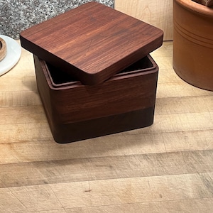 Peut inclure: Une boîte carrée en bois avec un couvercle, fabriquée en bois brun foncé. La boîte est ouverte, révélant un intérieur sombre. Le couvercle est légèrement entrouvert, mettant en valeur la construction de la boîte. La boîte repose sur une surface en bois.