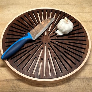 Puede incluir: Una tabla de cortar redonda de madera con un diseño de sol. La tabla tiene un anillo exterior de madera clara y un centro de madera oscura con ranuras cortadas en un patrón radial. Un cuchillo y un diente de ajo están sobre la tabla.