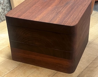 May include: A square wooden box with rounded corners, featuring a two-tiered design. The box is constructed from dark brown wood with a visible grain pattern, consisting of a lid and a base section that fit together to form a seamless cube shape.