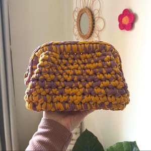 May include: A handmade crochet clutch bag in yellow and purple. The bag has a rounded shape and is held in a hand. The background includes a decorative mirror and a crochet flower.